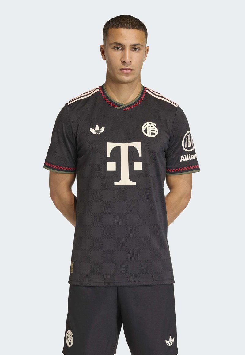 Mann steht mit Händen hinter dem Rücken, trägt ein schwarzes Fußballtrikot und Shorts mit weißen Logos und rot-grünem Saum am Kragen und den Ärmeln.
