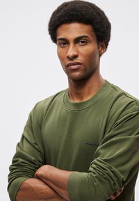 Sudadera verde oliva, cuello redondo, puños acanalados, tela suave, con un pequeño logo azul marino en el pecho, ajuste relajado, brazos cruzados.