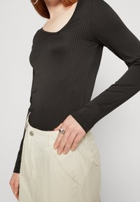 Černý žebrovaný top s dlouhými rukávy a kulatým výstřihem v kombinaci s světlými, texturovanými kalhotami s vysokým pasem, které mají kapsy a knoflíkové detaily.