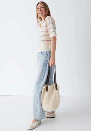 Lächelnde Frau in beigem gestreiftem Pullover und hellblauen Jeans hält eine gehäkelte Einkaufstasche mit dunklen Griffen vor weißem Hintergrund.