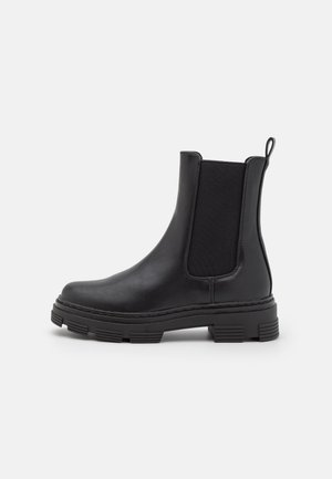 Botte Chelsea en cuir noir avec semelle en caoutchouc épaisse, panneaux latéraux élastiques et languette à l'arrière, vue de profil sur fond blanc.
