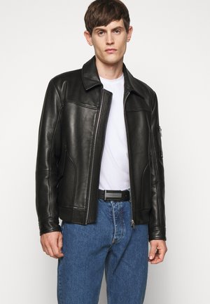 Chaqueta bomber de cuero negra con cremallera frontal, cuello de botones y bolsillos laterales. Lleva una camiseta blanca y se combina con pantalones de mezclilla azules.