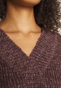 Strickpullover in einem tiefen Mauve-Ton mit einem V-Ausschnitt, der ein strukturiertes, grobes Maschenmuster aufweist. Weiches, warmes Material.