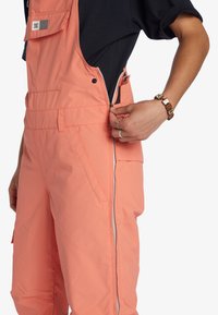 Roze, waterdichte, geïsoleerde overall met ritszakken aan de zijkant en aan de voorkant; verstelbare bandjes; zachte textuur.