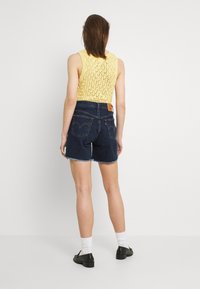 Jeanshorts med fransade kanter, tillsammans med en gul stickad linne. Baksidan har två fickor och en varumärkesetikett. Svarta skor fulländar looken.