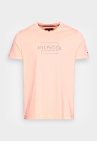 Camiseta de algodón color rosa claro con cuello redondo, que presenta un logo negro de "HILFIGER" en un diseño rectangular en el frente y mangas cortas.