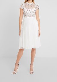 Valkoinen mekko, jossa kirjailtu kukkakuvio, tyllinen yläosa, hihat ja laskostettu hame. Yhdistetty hopeisiin remmikenkiin.