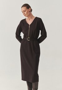 Zwarte jurk met een V-halslijn, lange mouwen en een ritssluiting aan de voorkant. Geverfde taille met een riem met een ronde gesp. Gladde textuur.