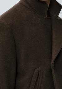 Cappotto in feltro marrone con colletto alto, caratterizzato da tasche angolari e una texture liscia. Il design mette in risalto una silhouette strutturata.