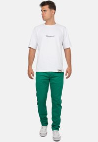 T-shirt bianca in cotone con logo nero in script, abbinata a pantaloni verdi e sneakers bianche. Design semplice con maniche corte.