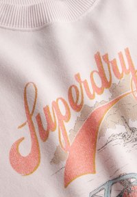 Lichtroze sweatshirt met retro-stijl "Superdry" tekst in oranje, geel en beige. Textuurstof met geribde kraagdetails.