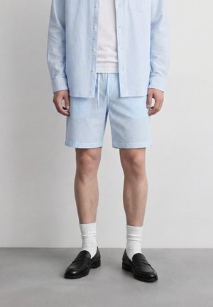 Homme portant un short à rayures bleu clair et une chemise à manches longues assortie, chaussettes blanches, mocassins noirs sur un sol gris devant un mur uni.