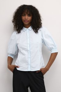 Blusa in cotone azzurro chiaro con maniche corte a sbuffo, apertura frontale con bottoni e pizzo lungo il colletto e il placket. Abbinata a pantaloni neri.
