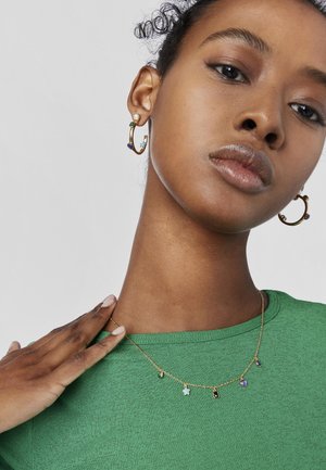 Collar de tono dorado con diversos charms que incluyen una estrella, gotas y piedras, llevado con una camiseta verde texturizada y aretes de aro.