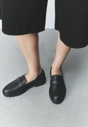 Benen met zwarte cropped broeken en zwarte leren loafers die op een grijze vloer staan.