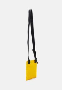 Borsa a tracolla rettangolare gialla con strap regolabile nera e chiusura superiore con zip, appesa su uno sfondo bianco.