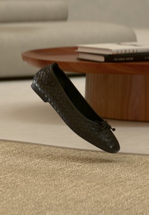 Zwarte gevlochten ballerinaschoen met een ronde neus, dunne zool en een klein strikdetail aan de voorkant. Geplaatst op een getextureerde beige ondergrond.