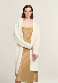 Langer weißer Cardigan aus strukturiertem, flauschigem Stoff, offene Vorderseite, mit weiten Ärmeln, getragen über einem glatten, goldenen Satin-Nachthemd.