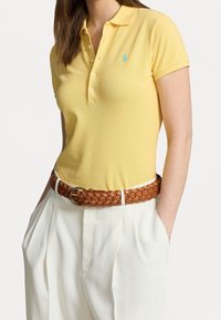Žluté polo tričko s klasickým límcem, krátkými rukávy a malým modrým logem. Spárováno s pleteným hnědým páskem a bílými kalhotami s vysokým pasem.