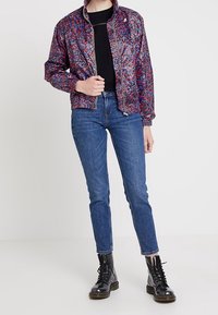 Giacca bomber multicolore con motivo floreale, cerniera frontale, T-shirt nera sotto, abbinata a jeans blu e stivali neri.