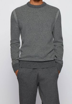 Man draagt een grijze slim-fit trui met lange mouwen en horizontale strepen en een bijpassende grijze broek, staand met handen in de zakken.