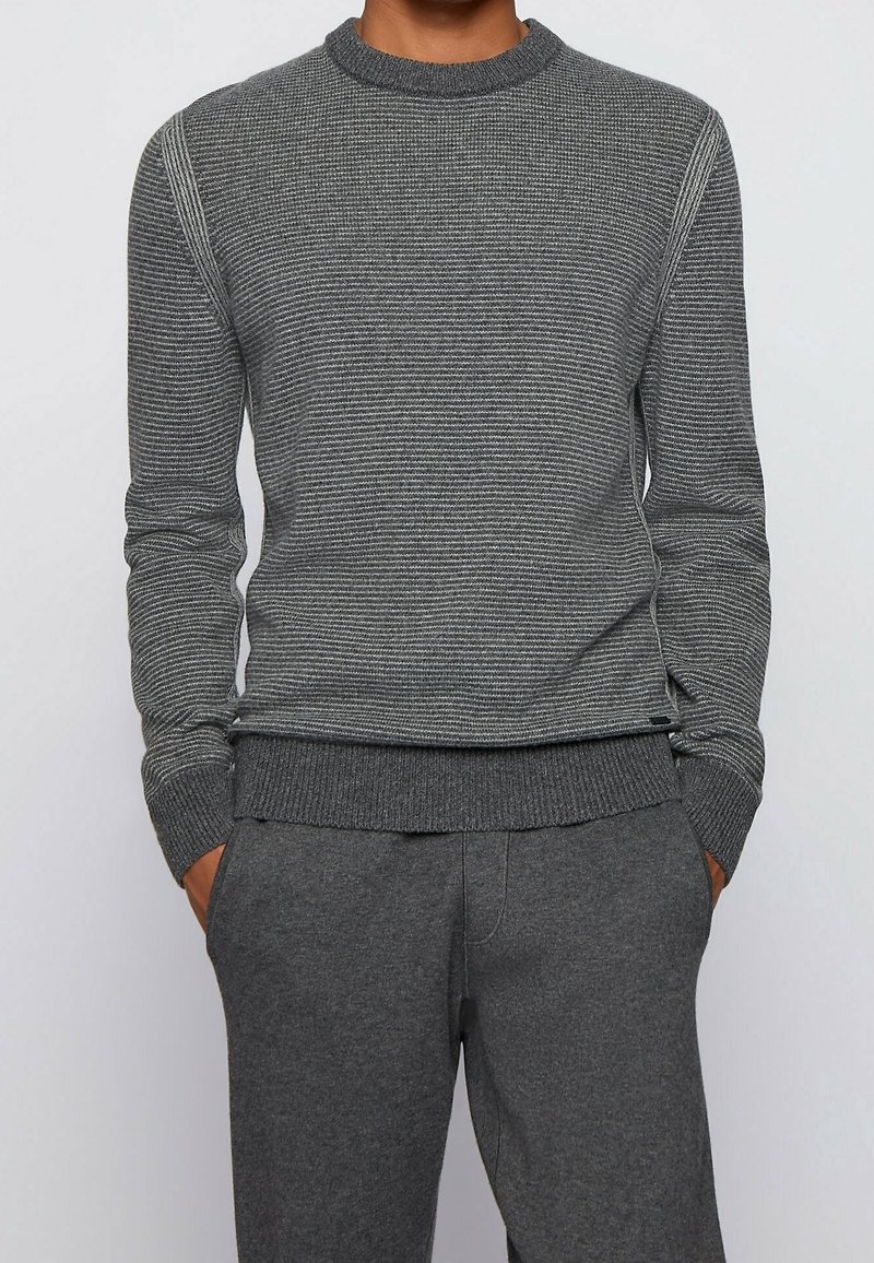 Homme portant un pull gris slim à manches longues avec des rayures horizontales et un pantalon gris assorti, debout les mains dans les poches.