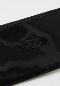 Cartera de charol negro con cierre de cremallera, que presenta una textura suave y un acabado brillante. Incluye un distintivo logo ovalado en el frente.