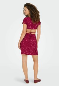Femme aux cheveux bouclés portant une robe à pois bordeaux à manches courtes avec le dos nu et des ballerines bordeaux, se tenant devant un fond uni.