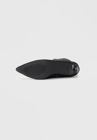 Stivale alla caviglia nero con punta appuntita mostrato con la suola verso l'alto, caratterizzato da un motivo di grip scanalato e un tacco a blocco basso su sfondo bianco.