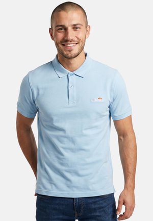 Homme souriant portant un polo ellesse bleu clair et un jean foncé, debout les mains derrière le dos sur un fond blanc uni.