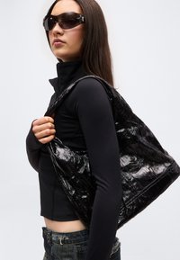 Νεαρή γυναίκα που φοράει μαύρη μπλούζα μακρυμάνικη, σκοτεινά γυαλιά ηλίου, κρατώντας μεγάλη γυαλιστερή μαύρη τσάντα στον ώμο, μπροστά σε απλό φόντο.