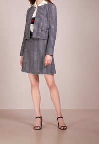 Giacca a quadri blu navy e bianca con design corto, chiusura con zip, abbinata a una gonna coordinata. La modella indossa tacchi neri con cinturino alla caviglia.