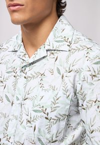 Camicia azzurra a maniche corte con un motivo di foglie verdi e marroni. Presenta un colletto e un singolo bottone sulla tasca del petto.