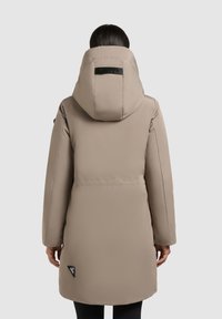 Person wearing a long beige padded coat with a large hood, shown from the back against a plain light background.