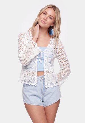 Giovane donna con capelli biondi che indossa un cardigan bianco all'uncinetto sopra un crop top azzurro chiaro e pantaloncini abbinati con orecchini di piume blu.