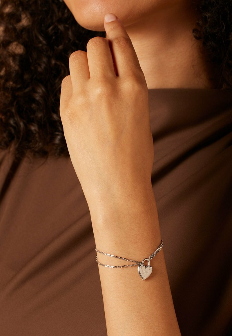 Zilveren armband met twee verbonden schakels, voorzien van een hartvormige hangslot charm. Glad oppervlak tegen een bruine stoffen achtergrond.