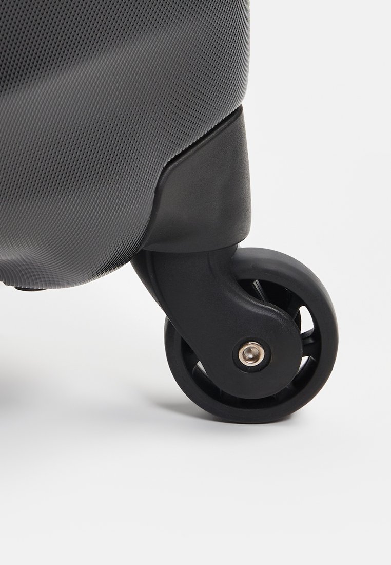 Coin de valise noire à roulettes avec un extérieur texturé. Équipée d'une roue noire solide avec un essieu métallique, conçue pour une mobilité facile.