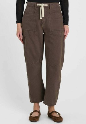 Pantaloni marroni dal taglio comodo con grandi tasche frontali e coulisse bianca in vita, indossati con scarpe basse marrone scuro e top nero a maniche lunghe.