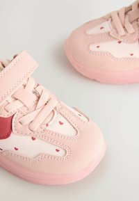 Baskets de sport roses avec une texture douce, dotées d'une sangle Velcro, de motifs en forme de cœur et d'accents rouges contrastants sur les côtés et les lacets.