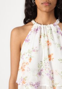 Hvid halterneck-top lavet af letvægtsstof, med flerfarvede blomsterprint i orange, lilla og grønt, og en samlet halsudskæring.