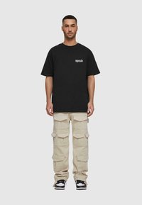 Sort T-shirt med korte ærmer og hvidt logo på brystet, parret med beige cargo bukser med flere lommer og en løs pasform. Sorte sneakers.