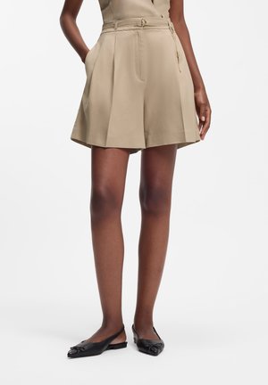 Femme portant un short plissé taille haute beige avec une ceinture et des ballerines à bouts pointus noires à bride arrière, debout avec une main dans la poche.