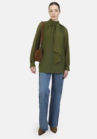 Long-sleeve, olive green blouse with a tie collar, light texture, paired with light blue wide-leg jeans and brown pointed-toe heels. Holds a brown bag.