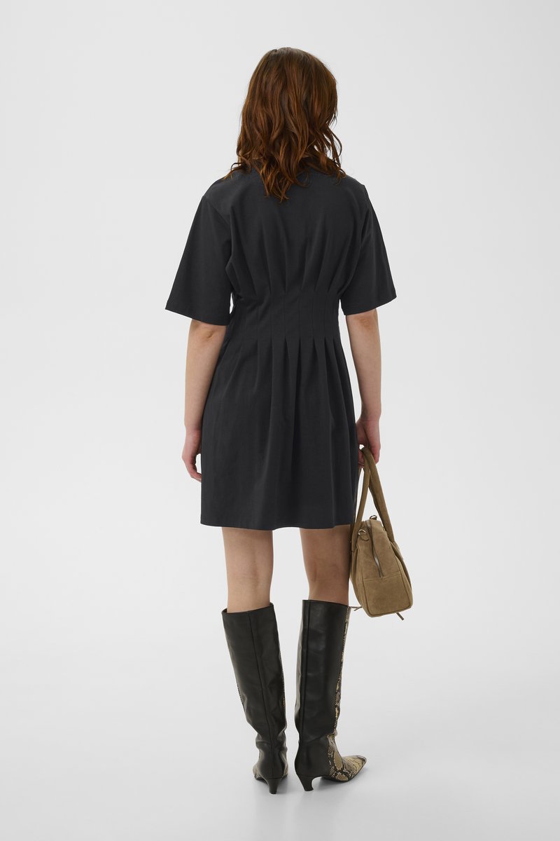 Femme aux cheveux bruns ondulés portant une robe courte noire, des bottes noires montantes à motif serpent, tenant un sac à main couleur tan, vue de dos.