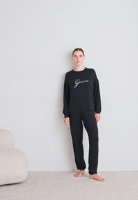 Eine Frau steht barfuß gegen eine helle Wand, trägt schwarze Jogginghosen und einen passenden schwarzen Sweatshirt mit "Guess"-Logo.