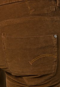 Bruine ribfluweelstof met horizontale ribbels, een achterzak die wordt benadrukt door gele stiksels en een metalen knoopdetail.