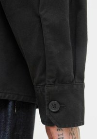 Black denim jacket sleeve with a buttoned cuff over a tattooed hand, showing fabric texture and stitching details.