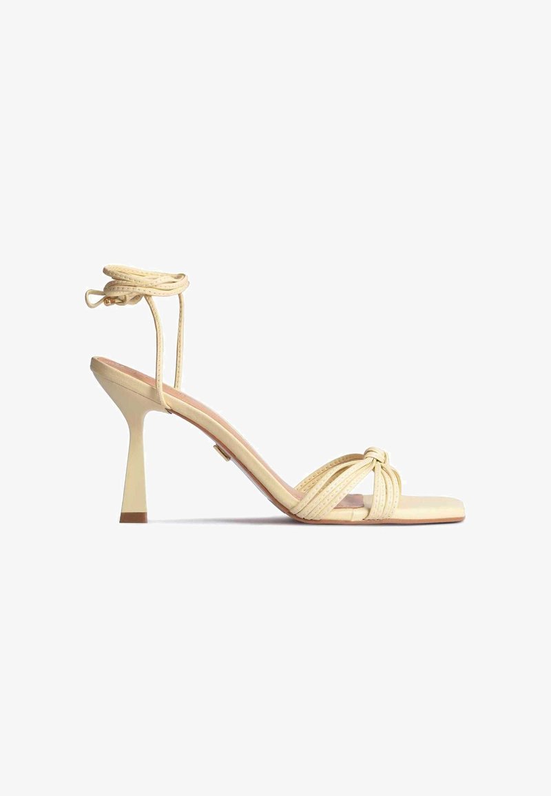 Sandale beige à talons hauts avec bout carré, lanières frontales torsadées et fines lanières à nouer autour de la cheville.
