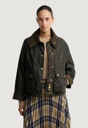 Femme portant une veste vert foncé avec un grand col par-dessus un pull beige et une jupe plissée à carreaux.