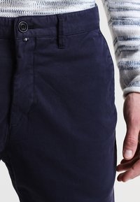 Granatowe spodnie wykonane z gładkiego bawełnianego materiału, z zapięciem na guzik, bocznymi kieszeniami oraz wyraźnymi detalami przeszyć w pasie.